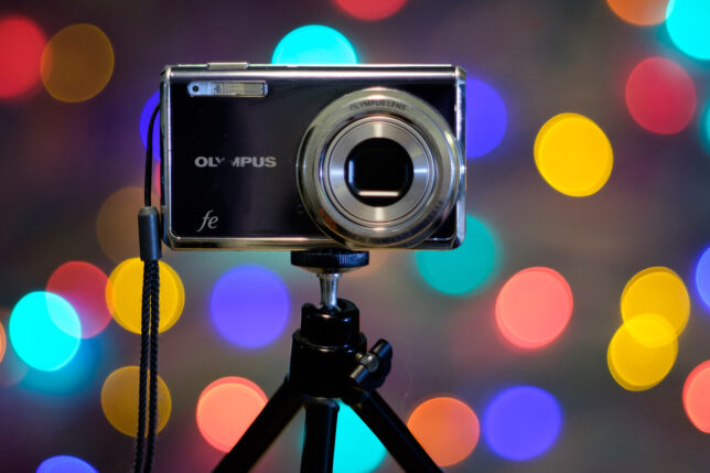 An old point-and-shoot camera sits on a tripod with Christmas lights in the background. 85mm Nikkor f/2.0 at f/2.0.